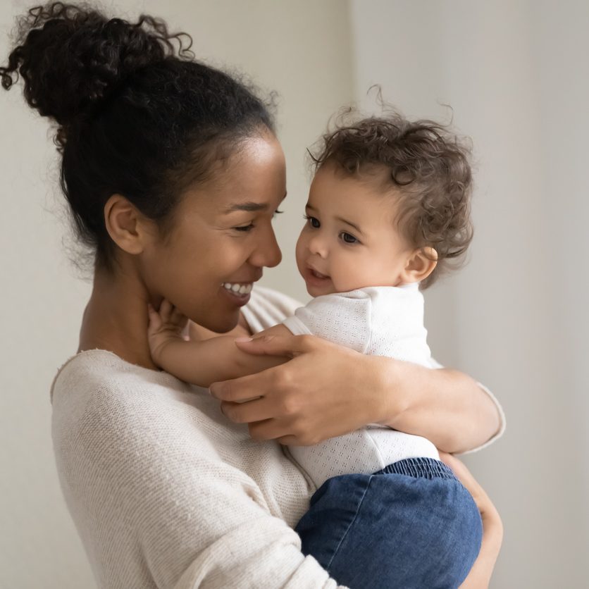 Head shot affectionate millennial biracial mommy or babysitter carrying on hands funny cute curly mixed race kid girl or boy, enjoying daycare time together indoors, responsible parenting concept.